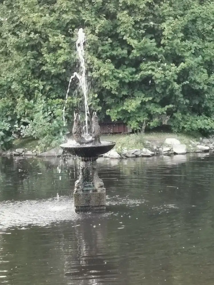 En lugn damm i sjöparkens camping med spegelblanka vattenytor och omgivande grönska, där en fontän syns i bakgrunden.