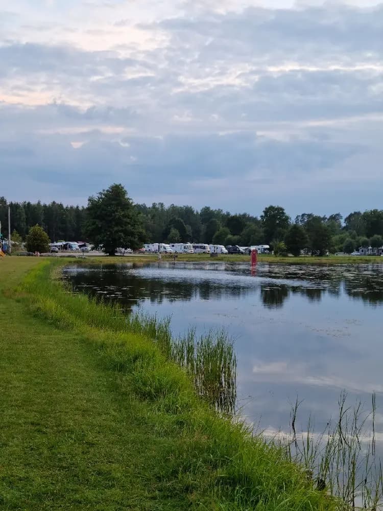 En lugn och stillsam sjö vid Skeppeviks camping, omgiven av vidsträckta ängar och våtmarker under en molnig himmel, med reflektioner i vattnet som skapar en harmonisk landskapsvy.