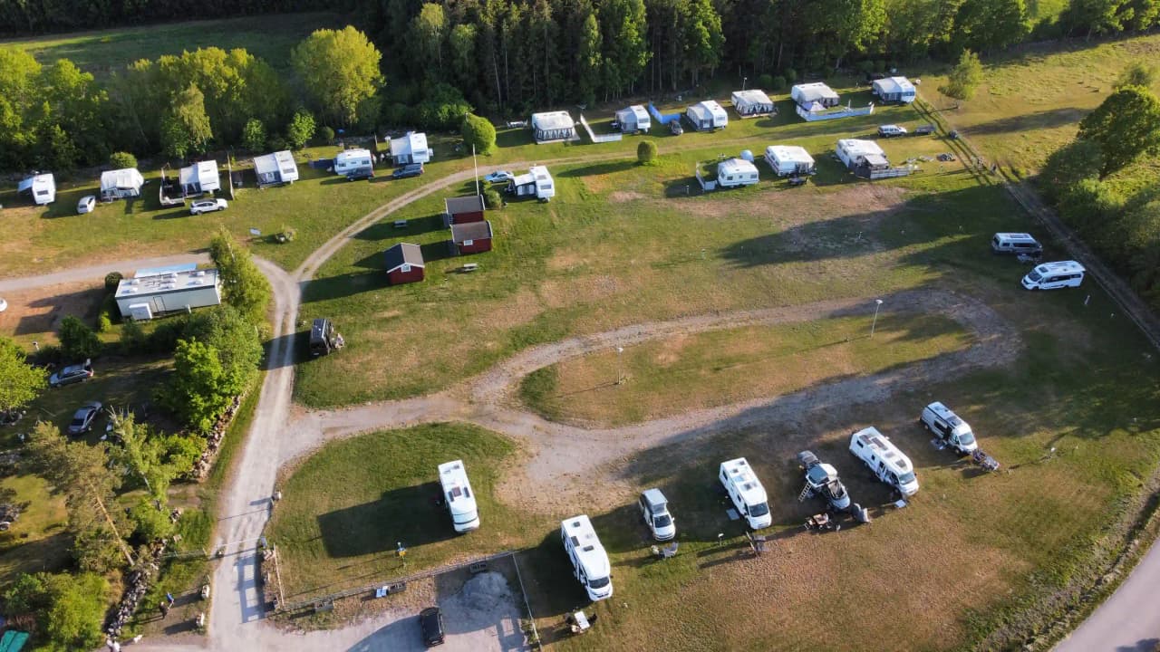 Fågelperspektiv av Skeppeviks Camping med bilar och husbilar parkerade på en öppen gräsplätt, omgiven av skog och natur.