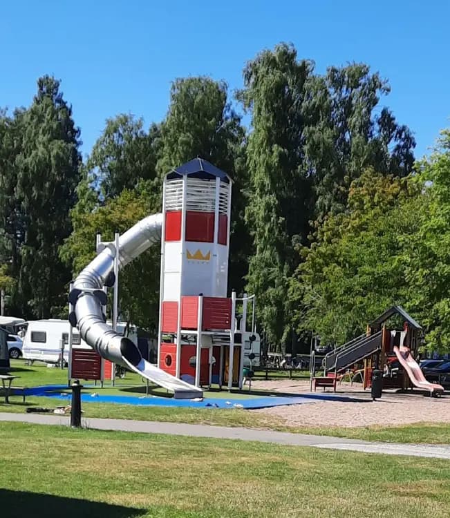 En lekplats med en rutschkana på Kronocamping, omgiven av gröna gräsmattor och skuggande träd i ett trevligt rekreationsområde.