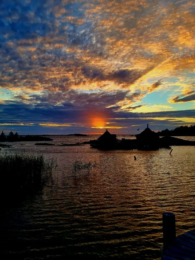 En vacker solnedgång över Filsbäcks camping med himlen färgad i varma toner. Lugnt vatten reflekterar himlens färger medan horisonten suddas ut vid skymningen. Moln drar försiktigt över himlen, vilket skapar en rofylld och naturskön atmosfär.