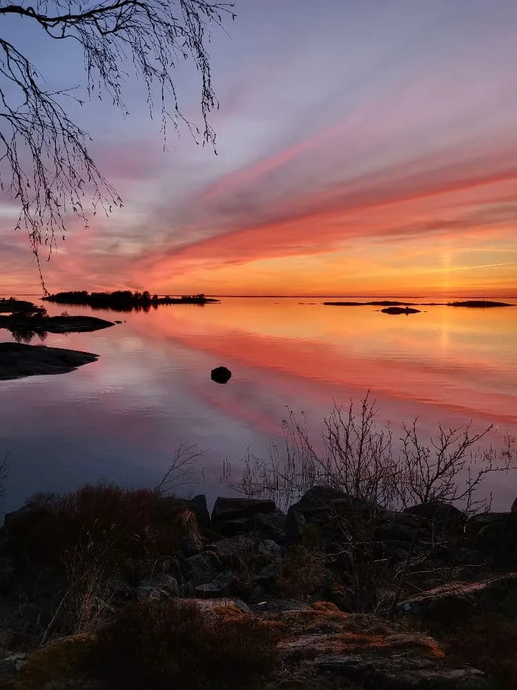 En solnedgång över horisonten vid Filsbäcks camping, där himlen skiftar i orange och rosa toner. Molnen speglas i det stilla vattnet och skapar en lugn och naturskön atmosfär.