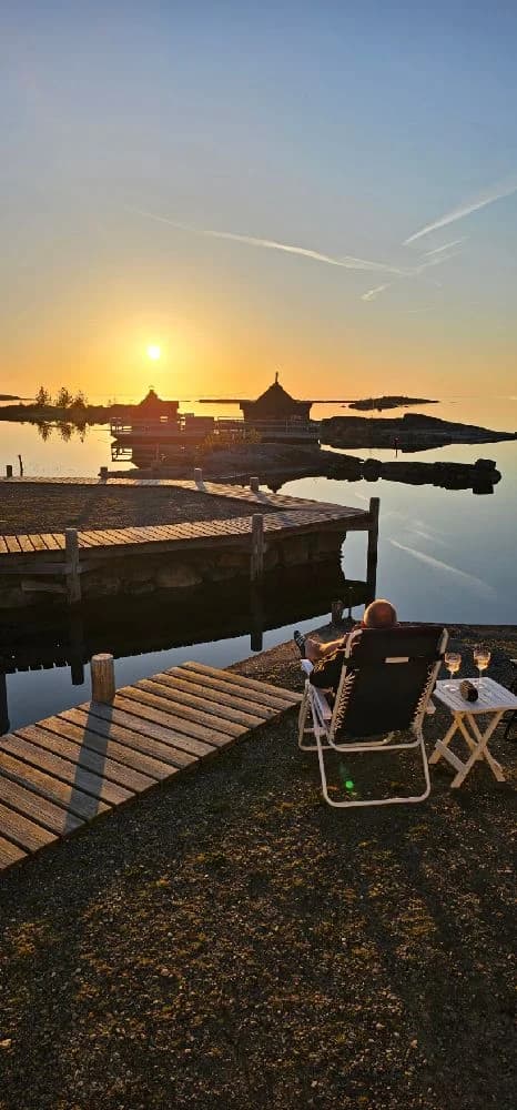 En stämningsfull solnedgång vid Filsbäcks camping med vattnet i förgrunden, himlen färgas i varma toner av gult och orange och speglas mjukt i det lugna vattnet.