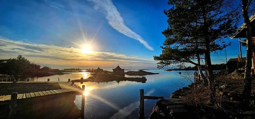 En vacker solnedgång vid Filsbäcks camping med vy över det klarblå vattnet som speglas i horisonten. Stenar kantar stranden under den dämpade skymningen.