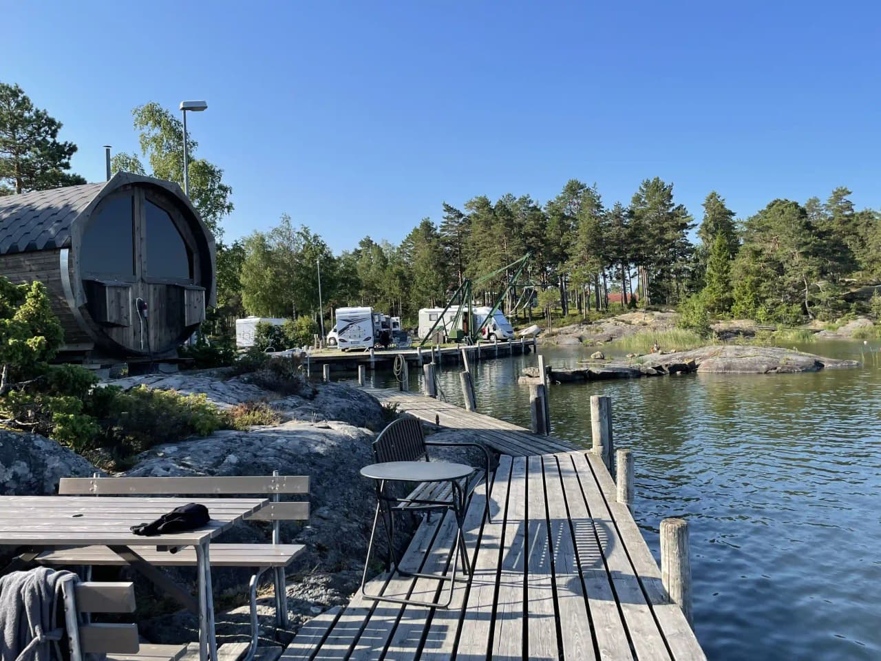 En vy över Filsbäcks camping med sjö och strand i förgrunden, där en brygga sträcker sig ut över vattnet. På den avlägsna stranden syns stugor omgivna av grönska.