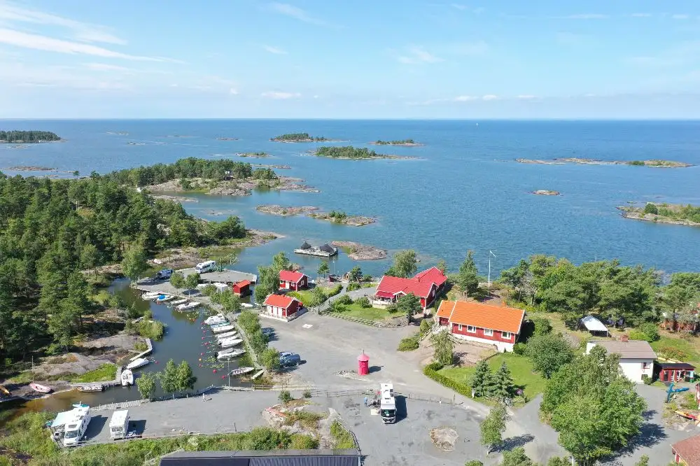 Flygbild över Filsbäcks camping som visar en kuststräcka med strand och vatten, samt båtar vid småbåtshamn. I bakgrunden syns en resortliknande miljö med grönska och byggnader.