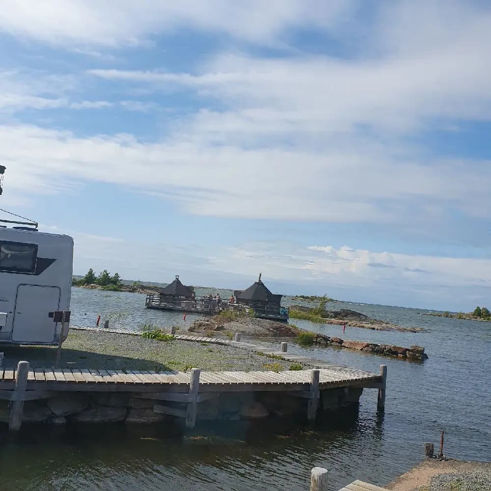 En vy över Filsbäcks camping med en vattenresurs i förgrunden. Flera båtar ligger förtöjda längs med strandkanten, och man kan se både vattenvägar och kustlandskap i bakgrunden.