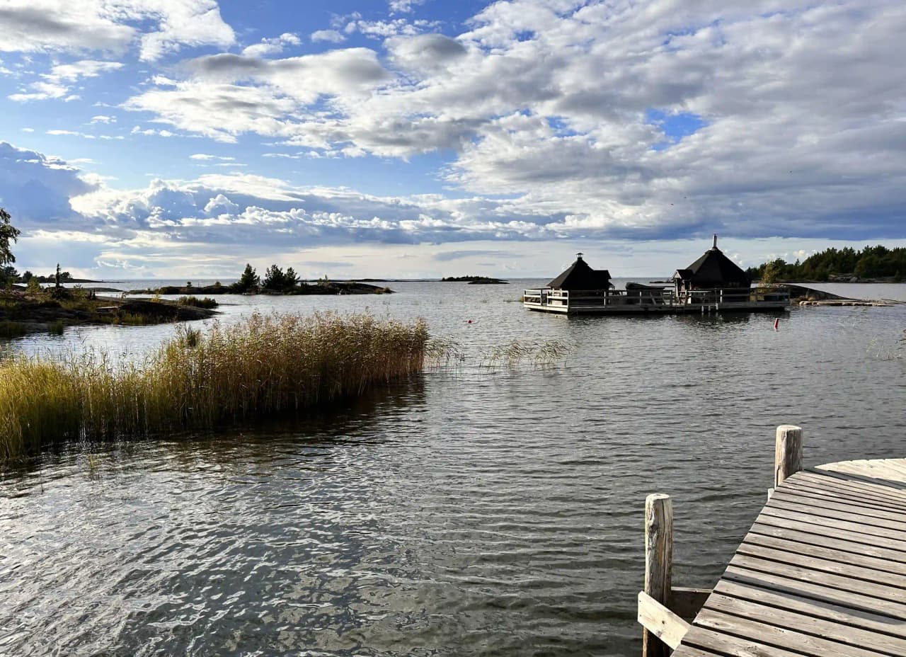 En naturskön vy över Filsbäcks camping med vattenspegel från en närliggande sjö, omgiven av grönskande bankar och en lugn kanal som återspeglar den omgivande naturens skönhet.