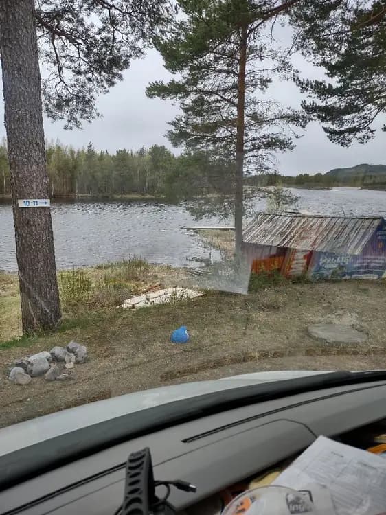 En kompaktbil står parkerad vid en glittrande sjö i ett naturskönt område med böljande kullar i bakgrunden, vilket framkallar en känsla av en idyllisk road trip vid Sonfjällscampen.