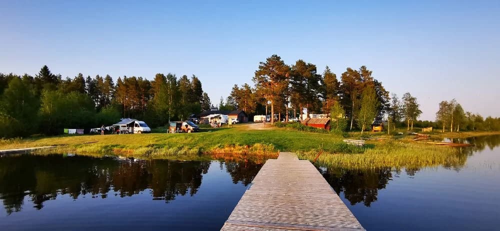 En somrig vy vid Sonfjällscampen med frodiga träd och växtlighet som omger en stilla sjö. Vattnets blanka yta speglar landskapet och förstärker den natursköna miljön.