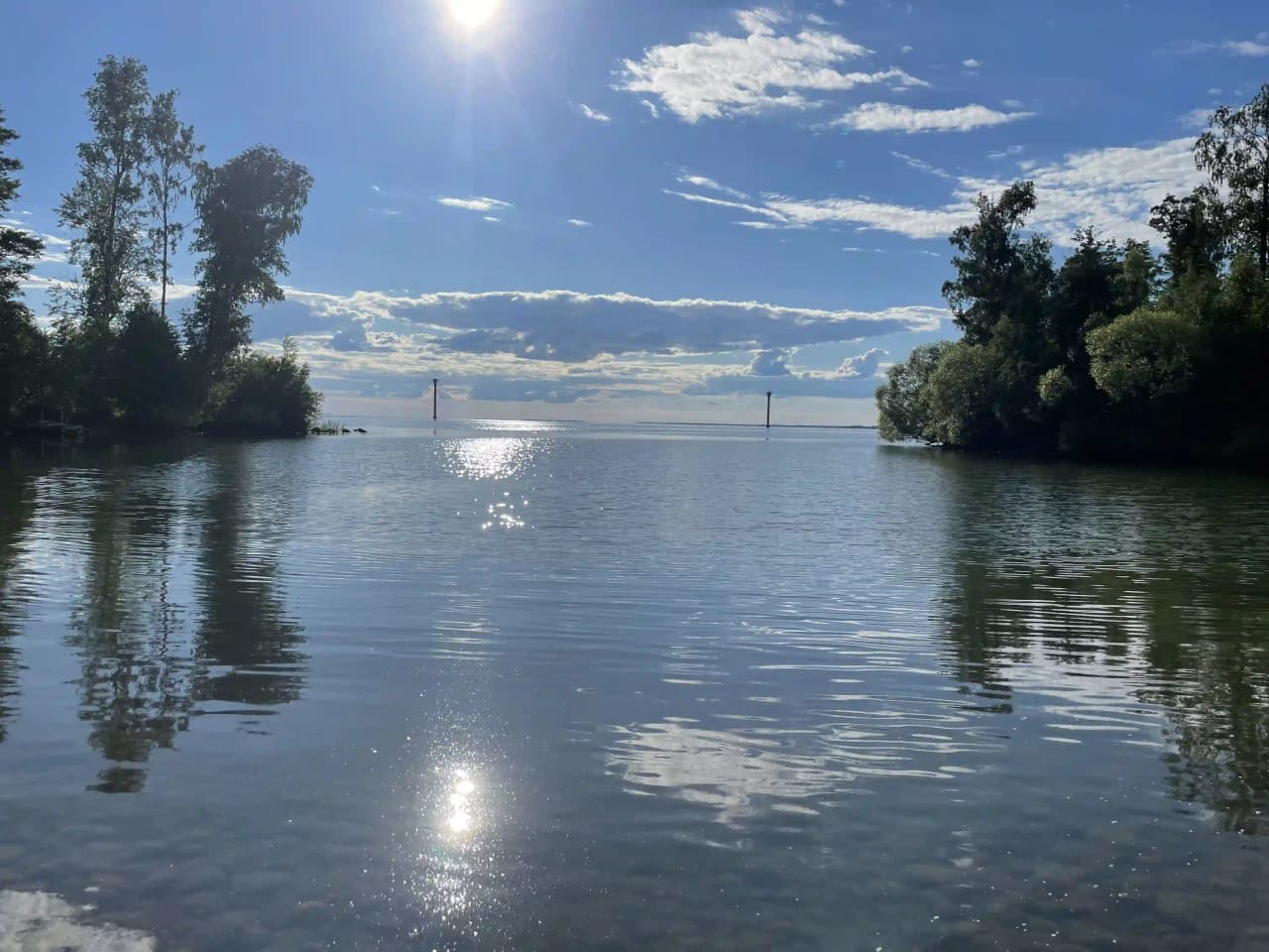 En solig dag vid Hjälmaresunds camping med utsikt över den blå sjön. Vattnet speglar de lätta molnen på himlen, och stränderna kantas av grönska.