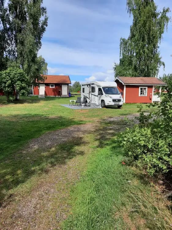 En familjebil parkerad nära en stuga på Hjälmaresunds camping, omgiven av grönska och gräsmatta. En bils bakljus syns, och stugan ligger i en trädgårdsliknande miljö.