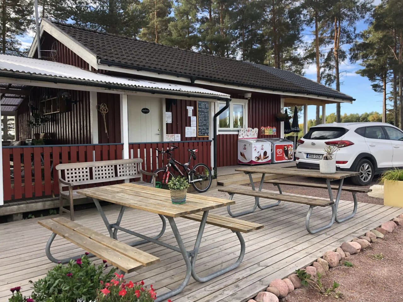 En vit stuga med veranda vid Våmåbadets camping, omgiven av träd och en grönskande trädgård i skuggan.