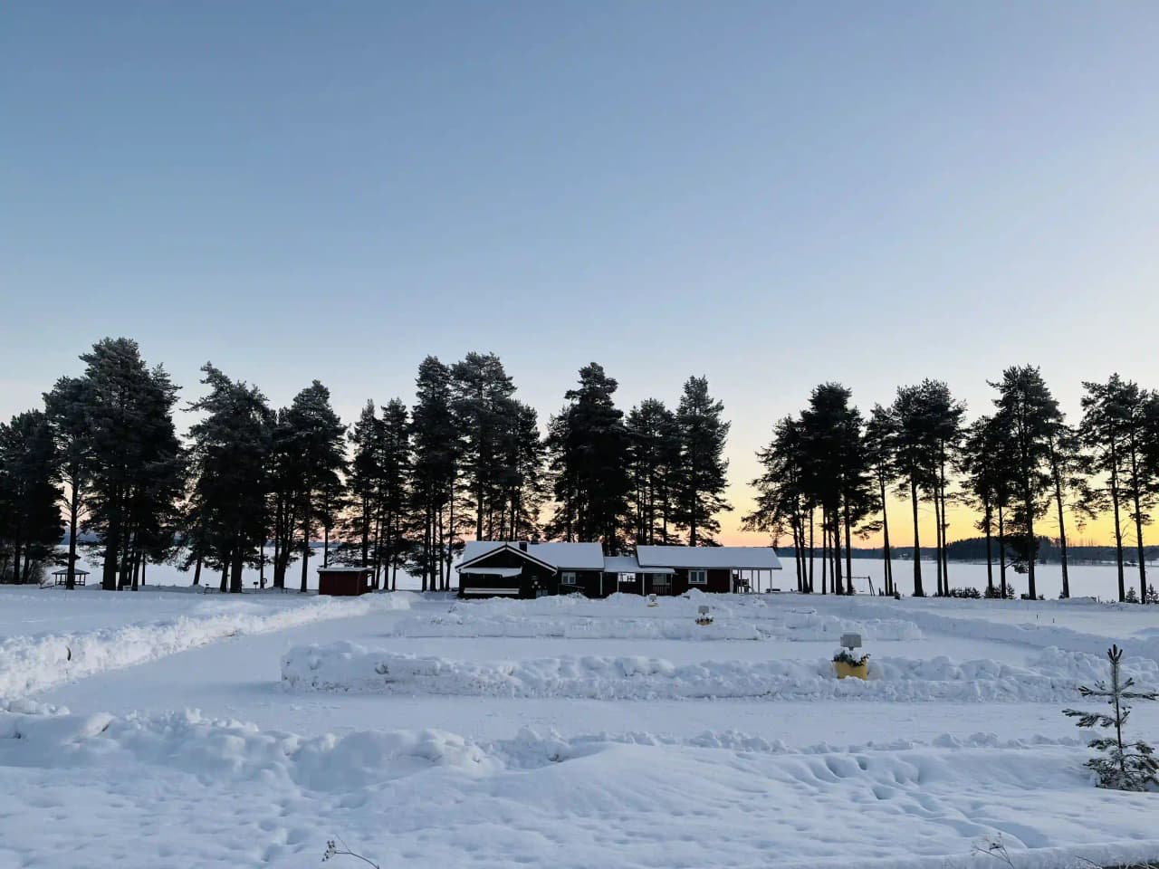 Vy över Våmåbadets camping under vintern. Snötäckt landskap med solens strålar som lyser genom morgondiset. Trädgrenar täckta av snö inramar bilden och sluttningar syns i bakgrunden.