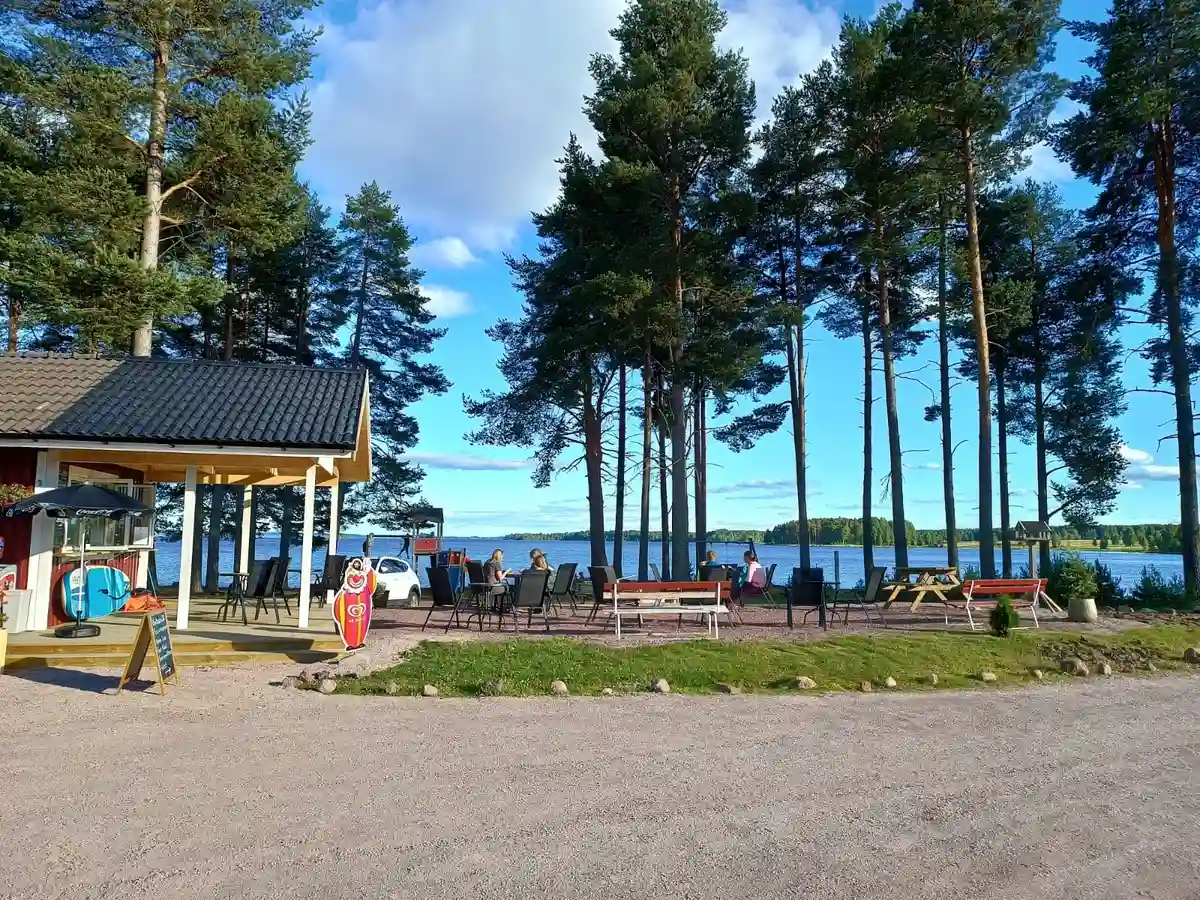 En fridfull kustlinje vid Våmåbadets camping med en sjö i bakgrunden, omgiven av skog och mjuka sandstränder. Ett par solskyddsstolpar finns placerade vid stranden, vilket ger en skuggig plats för besökare.