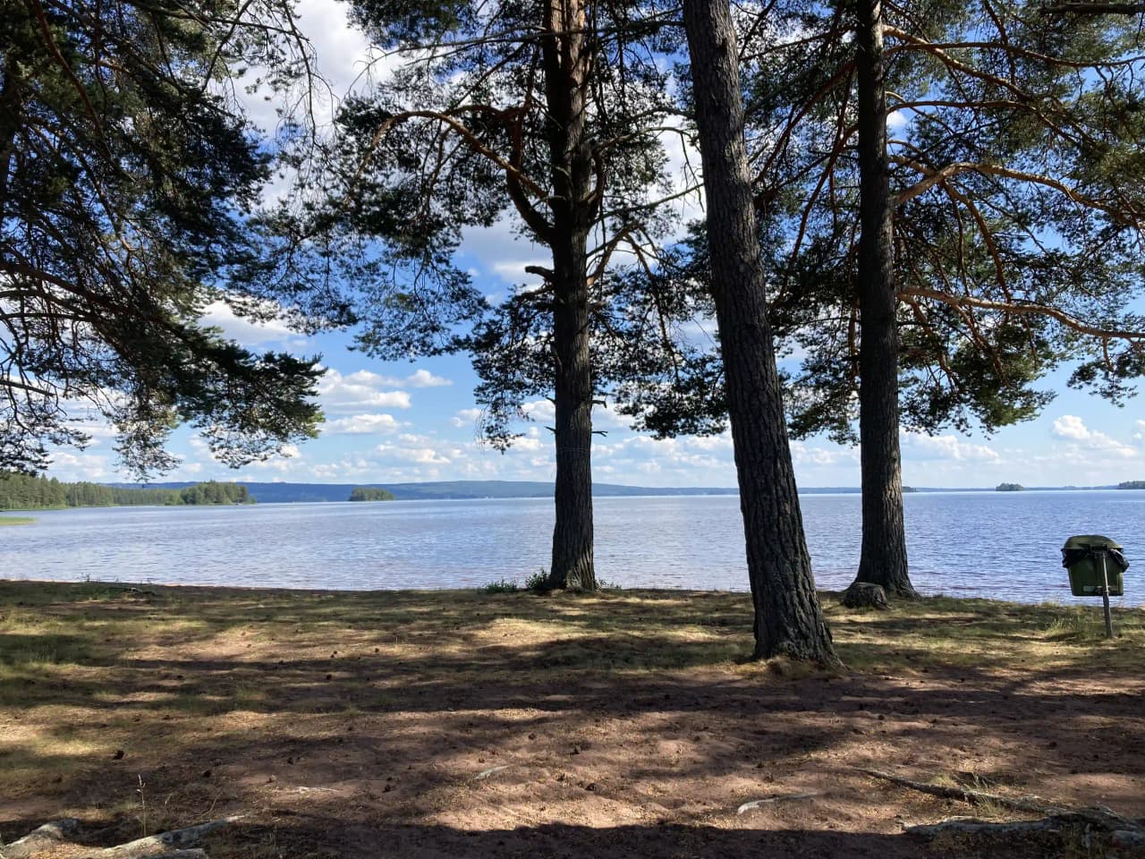 En naturskön vy över våmåbadets camping med vatten i förgrunden och en klarblå himmel i bakgrunden. Gröna trädgrenar ramar in landskapet vid vattenkanten.