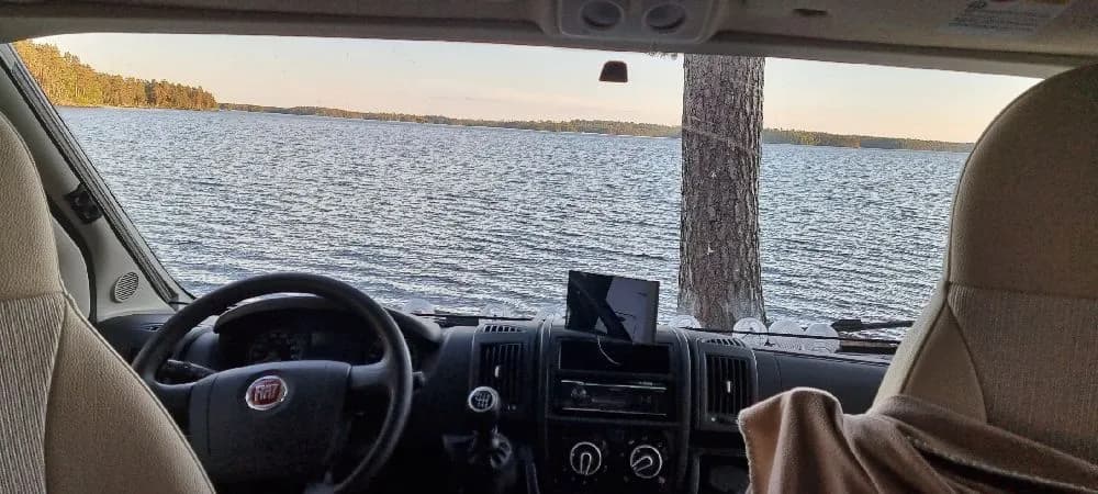 En minivan står parkerad vid Revelbadets camping, omgiven av grönska, med campingutrustning synlig genom bilfönstret.