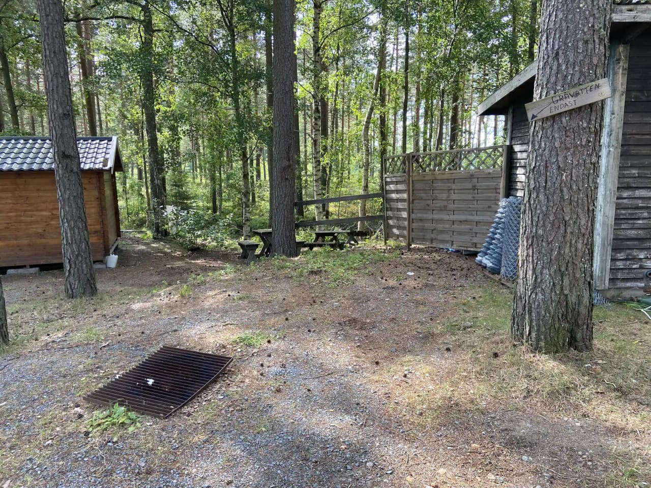 Bild av Revelbadets camping, visar en grön och lummig campingplats med skog och träd i bakgrunden. I förgrunden syns ett träd och ett mindre träskjul som ligger i ett naturskönt, lantligt område.