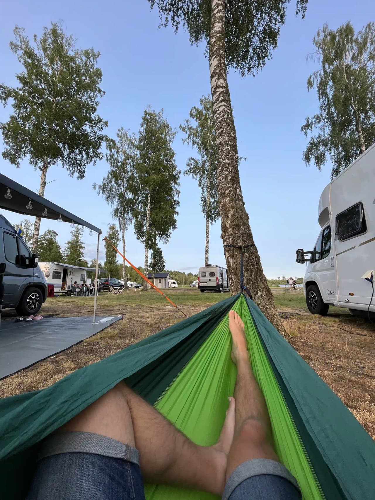 Personer som kopplar av i naturen på Hultsfred Strandcamping, omgiven av tält och hängmattor under skuggbildande tarpar.
