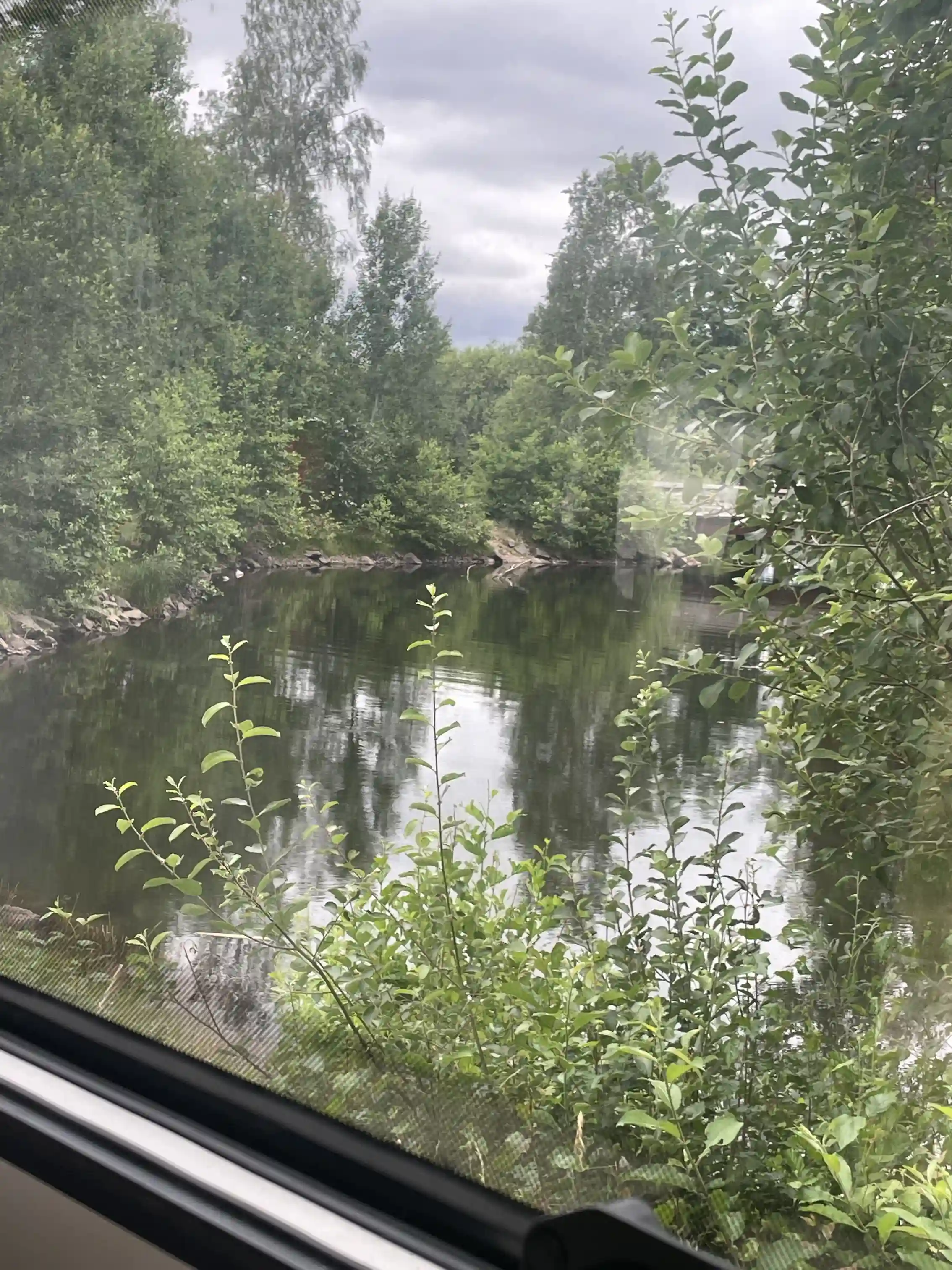 En naturskön vy vid Hultsfred strandcamping med en lugn vattenväg omgiven av skog och flodbankar, där träden speglas i det stilla vattnet.