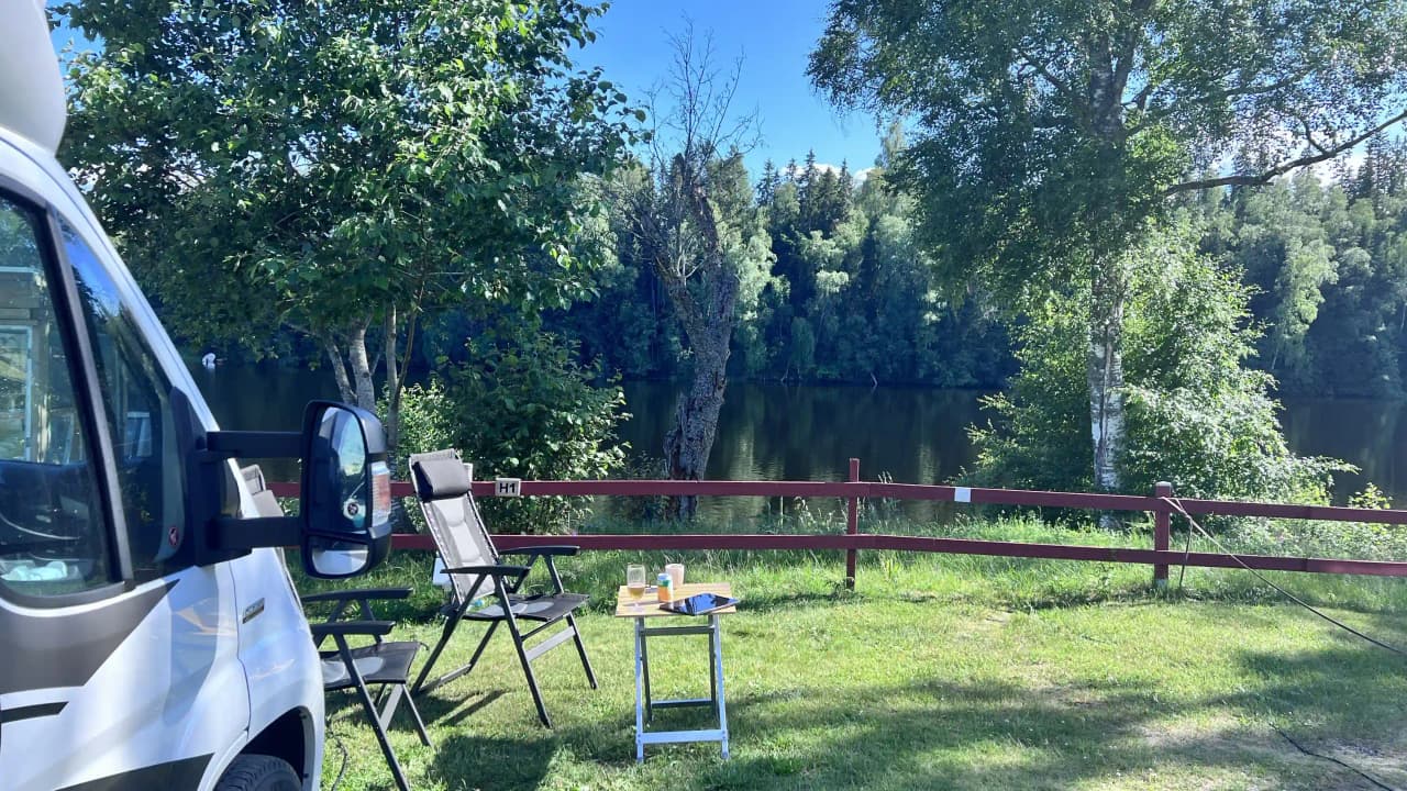En picknickplats med ett bord och bänkar på en gräsmatta, omgiven av frodigt grönområde. I bakgrunden syns en vit skåpbil parkerad, allt under en klarblå himmel.