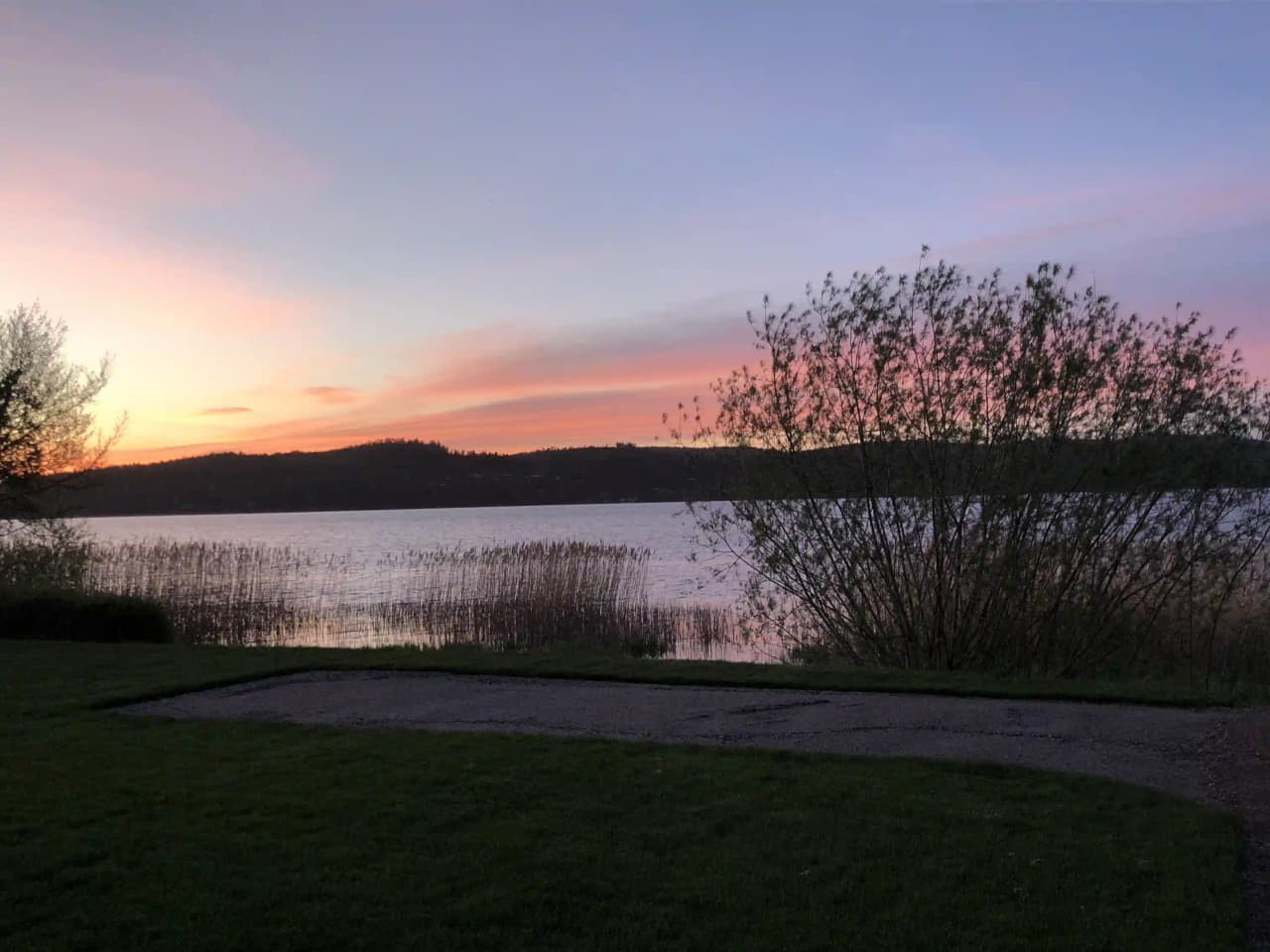 Vy över Lövekulle camping med ett stilla vattendrag i förgrunden, omgiven av grönska. Solnedgångens varma sken färgar horisonten och skapar en lugn och vacker atmosfär vid campingområdet.