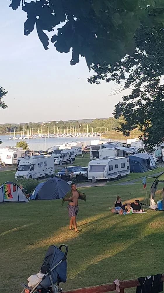 En campingplats med tält och husvagnar nära kusten, med båtar i bakgrunden.