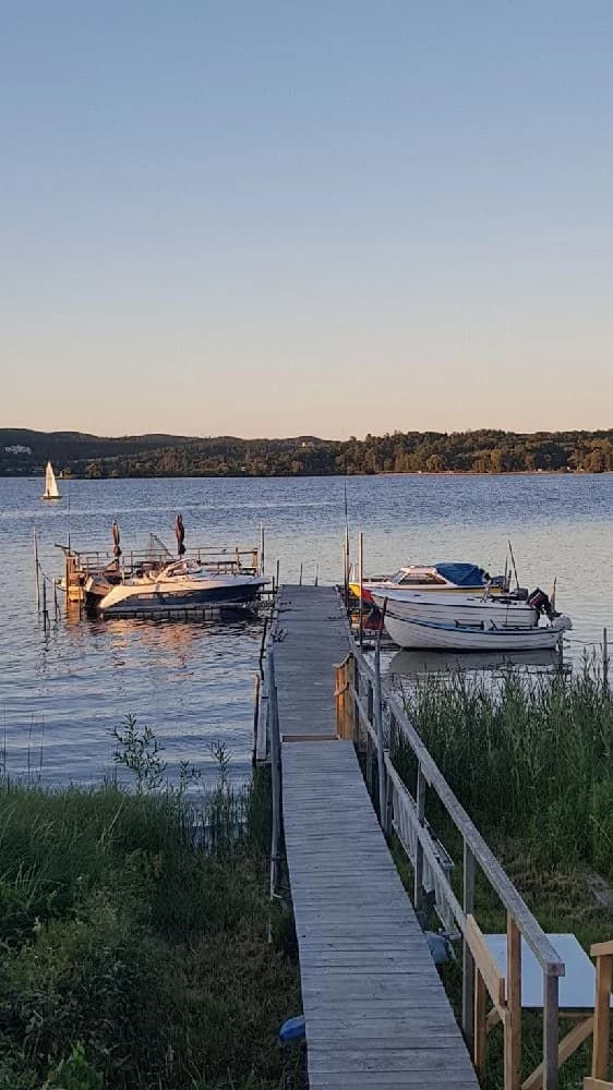 En vy över Lövekulle Camping med en sjö i förgrunden där flera båtar ligger vid en brygga. Området är omgivet av grönska och skog, vilket skapar en naturskön miljö.