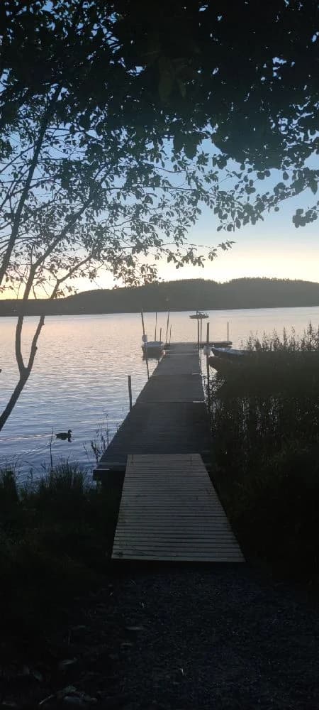 En stilla sjö vid skymning, med en enkel brygga som sträcker sig ut över vattnet. Horisonten syns svagt i bakgrunden, och omgivningen präglas av naturens lugn.