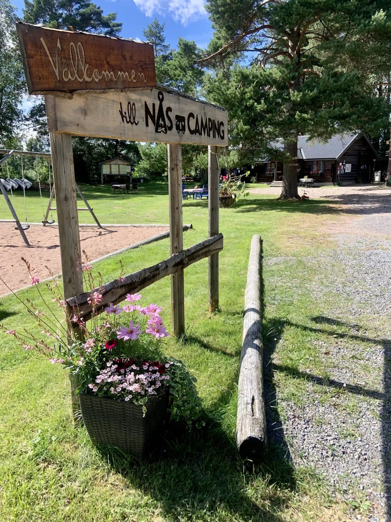 En grönskande campingplats med träd som ger skugga, en skylt som markerar området, och en välskött gård som skapar en inbjudande atmosfär för besökare.