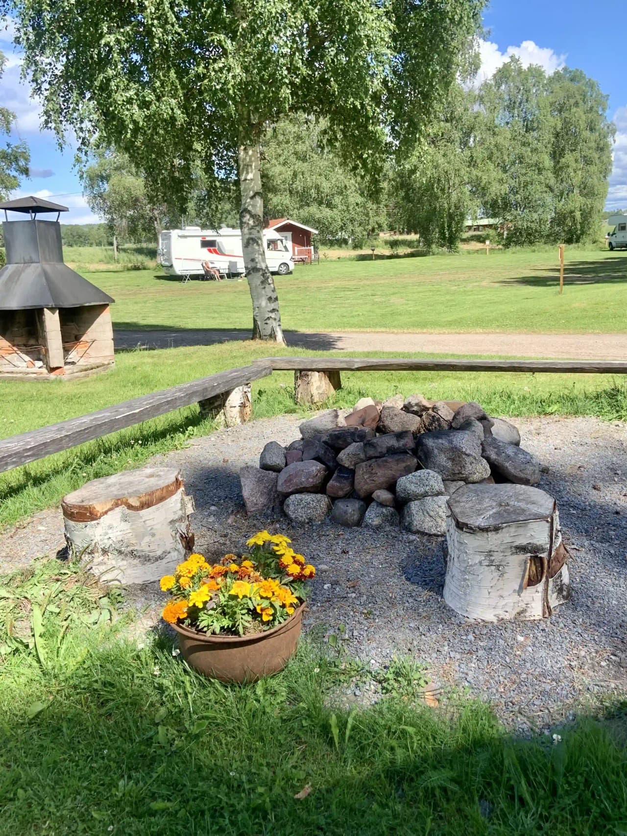 En campingplats med grönskande trädgård, gräsmatta och flera utomhusmöbler. Det finns träd som ger skugga och en stenbelagd yta.
