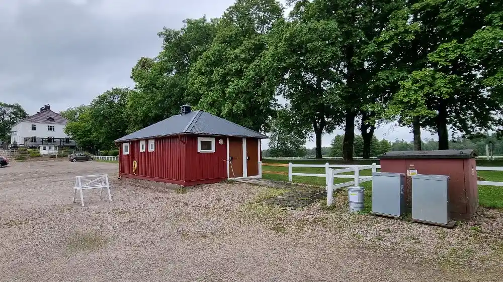 En pittoresk herrgård med tegeltak belägen i ett lantligt område. Byggnaden omges av en välskött gräsmatta och det finns en lada eller uthus i bakgrunden, vilket bidrar till en idyllisk och lantlig atmosfär.