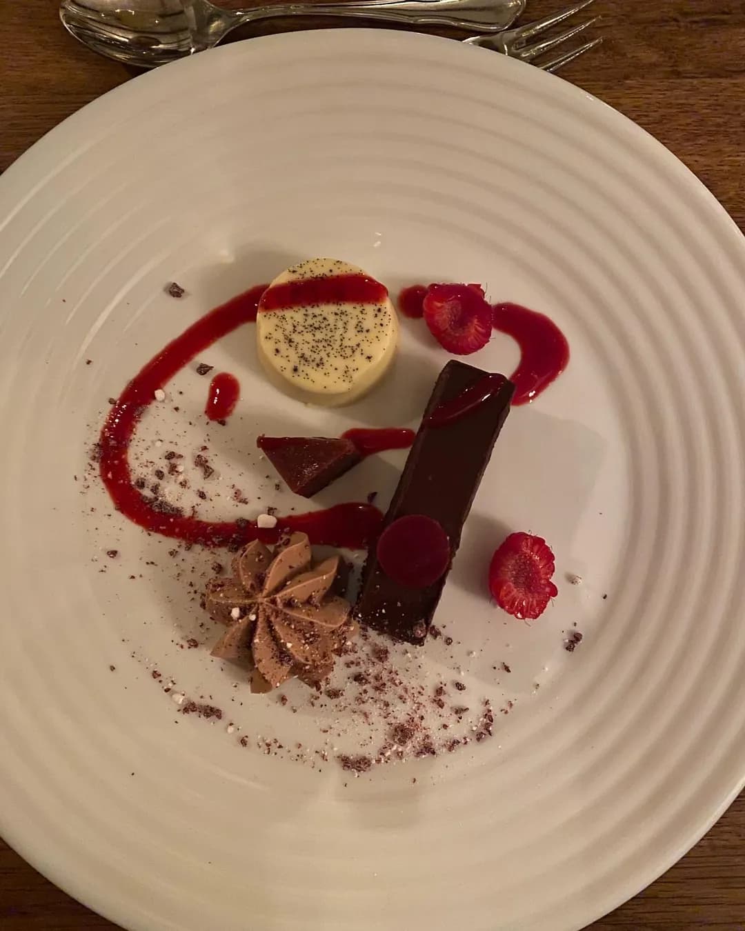 En elegant upplagd dessert på en vit tallrik, garnerad med färska bär och en grön ört, placerad på ett rustikt träbord. Inramningen antyder en mysig och inbjudande stämning, lämplig för en kulinarisk upplevelse på Hotell & Spa Lögnäs Gård.
