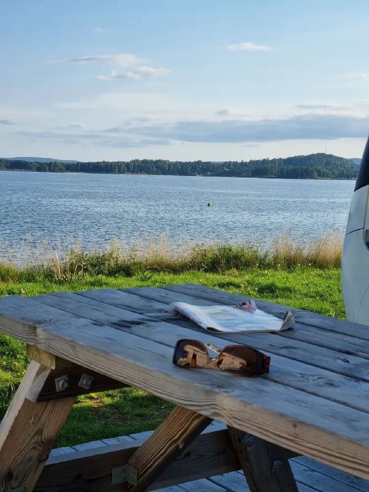En bild av Arvika småbåtshamn med picknickbord och bänkar i förgrunden. Solen skiner över sjön och hamnens kustlinje, medan moln syns på himlen i bakgrunden.