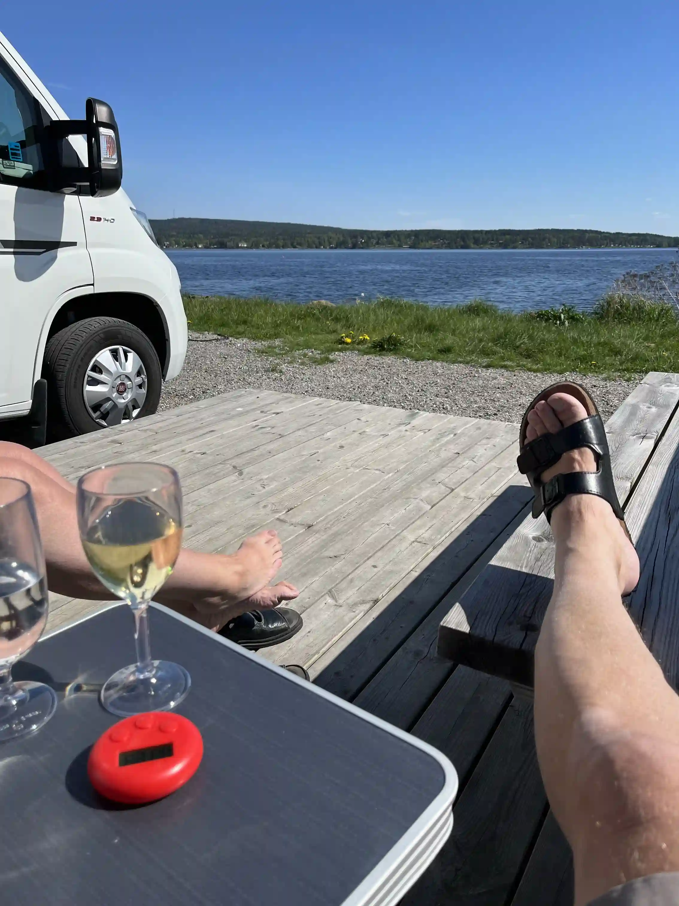 En solig dag vid Arvika småbåtshamn med människor som njuter av fritid och rekreation. I förgrunden syns ett utebord med flaskor, i bakgrunden finns båtar och husbilar.