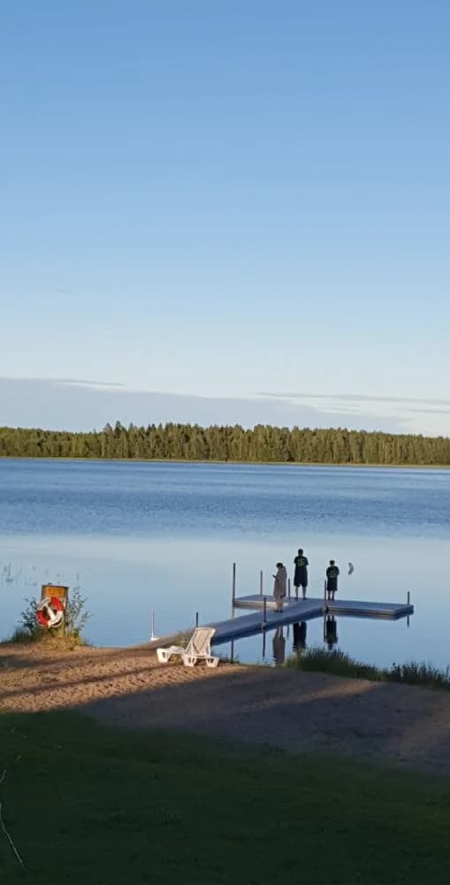 En fridfull vy över Mosjöns Camping, med blå himmel och en stor sjö i förgrunden omgiven av grönskande strandkant. Perfekt för rekreation och avkoppling vid vattnet.