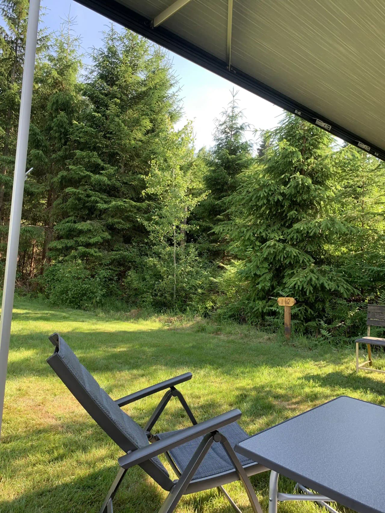 En grönskande campingplats med utemöbler under skuggande träd, omgiven av välskött gräsmatta och trädgård, med en avslappnad atmosfär i en park- eller trädgårdsmiljö.