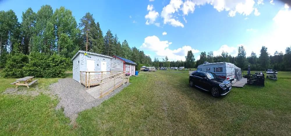 En husvagn och en husbil är parkerade i en grönskande campingmiljö med gräsmattor och höga träd. I bakgrunden syns fler fordon och området omges av skog.