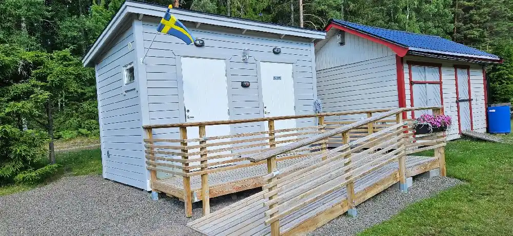 En trästuga med sidoklädsel av plankor och en liten veranda med trappsteg. Stugan omges av grönska och används som boende på Svennevads camping.