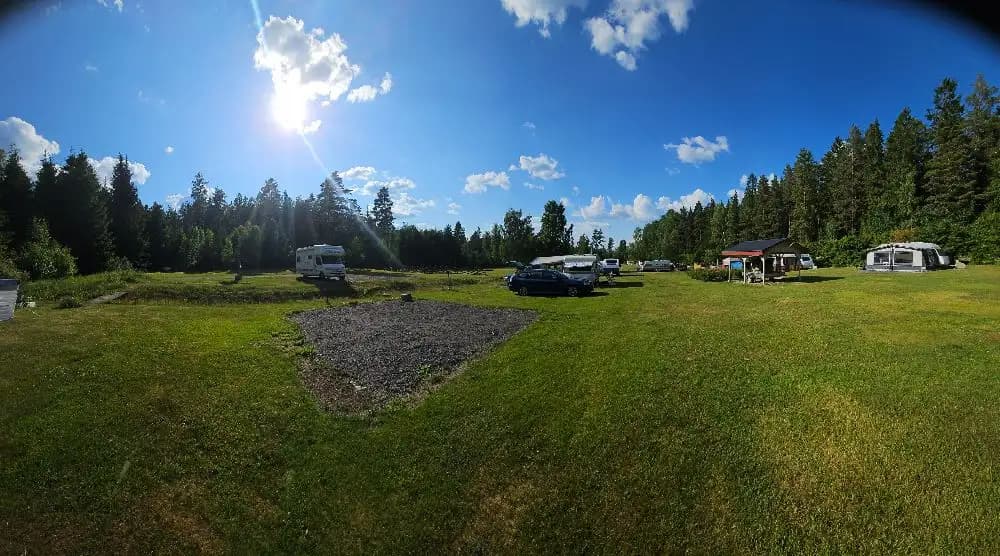 En grönskande campingplats med tält uppställda på en gräsmatta omgiven av lummiga träd och en klarblå himmel med vita moln i bakgrunden.