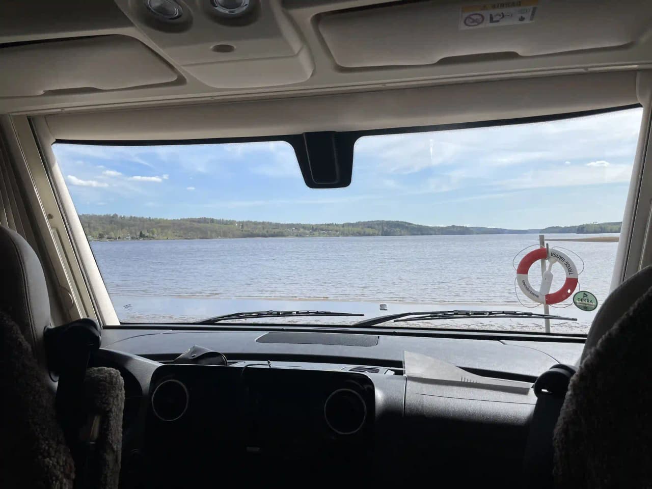 Utsikt från en bilkabin där förarens perspektiv visar en klar bilruta och sidospegel, med vägen framför som leder mot Hanatorps Camping, omgiven av en vidsträckt ocean i bakgrunden.