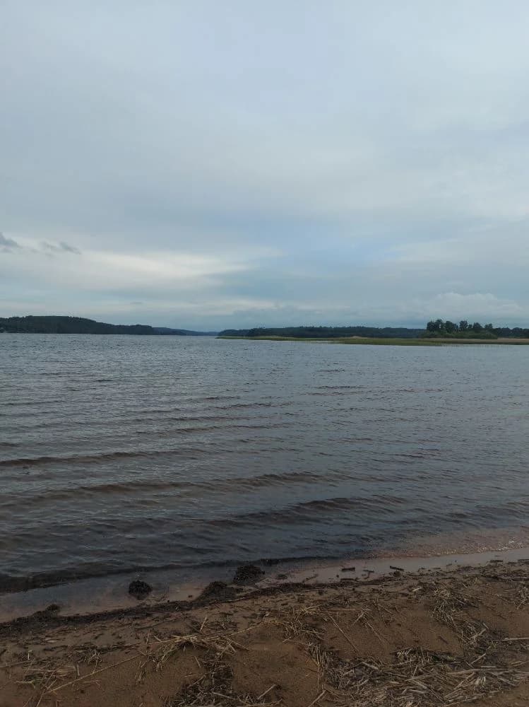 Utsikt över Hanatorps camping med klarblå himmel och molntäckt horisont, en vidsträckt vattenyta sträcker sig mot en strand och omgivande kustlandskap.