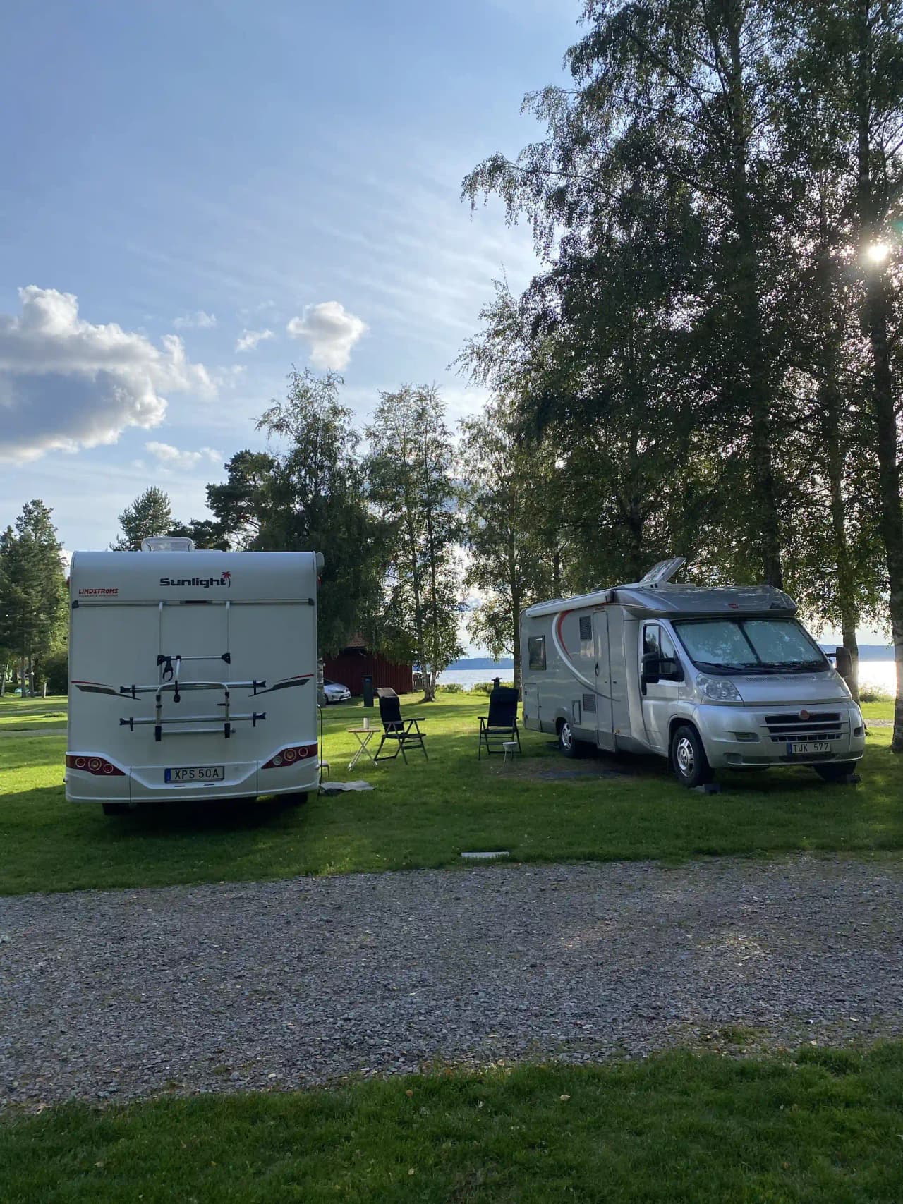 En grön campingplats med flera husvagnar och husbilar parkerade på gräsmattan. I bakgrunden syns träd och en klar himmel. Besökarna har ställt upp fordonen i en rymlig och välordnad miljö för en trevlig campingupplevelse.