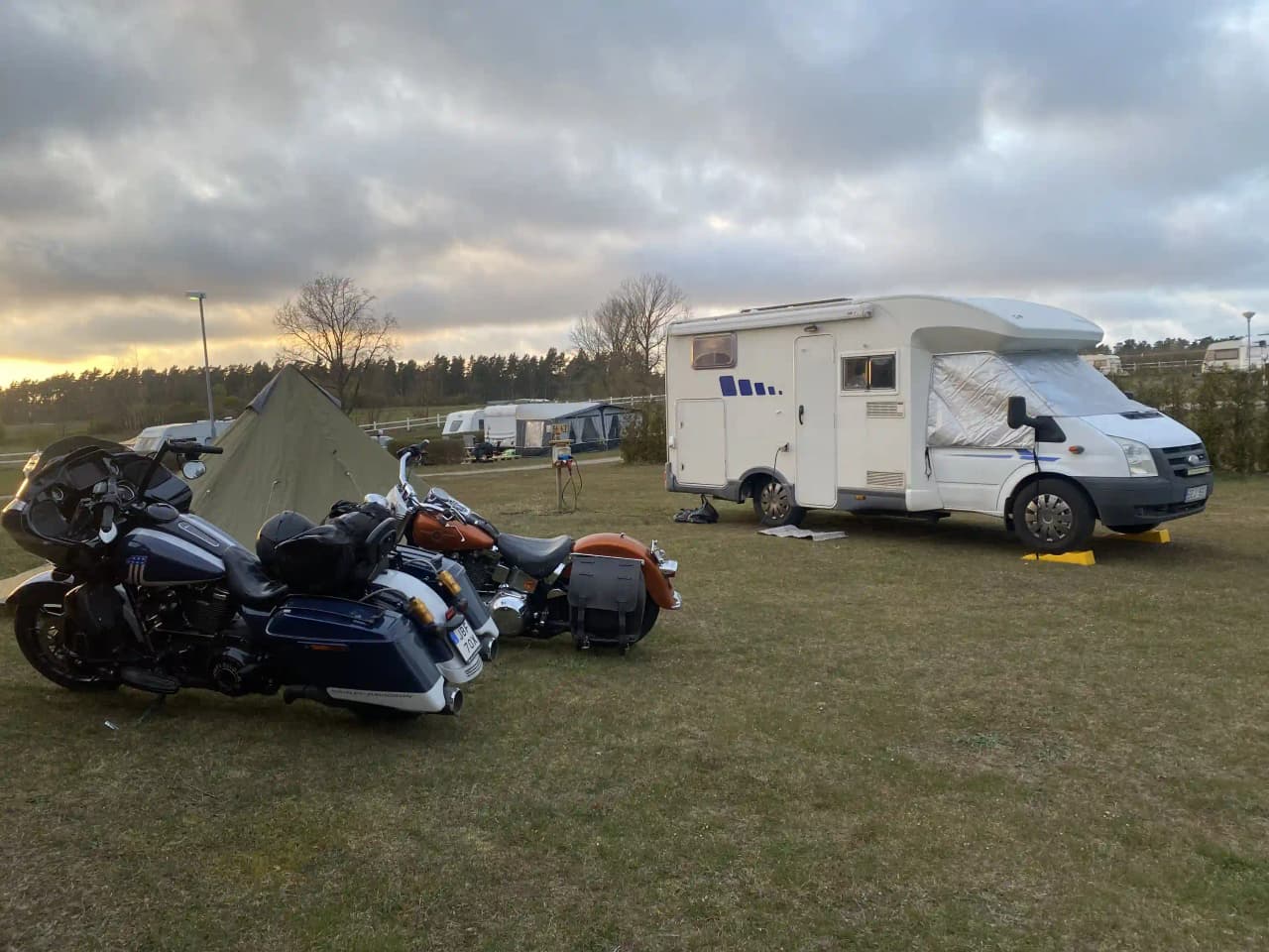 En husbil parkerad på ängdala camping, med fokus på fordonets utsida och hjul. I bakgrunden syns gröna träd och en klar himmel, vilket förstärker känslan av en naturskön campingmiljö på Österlen.