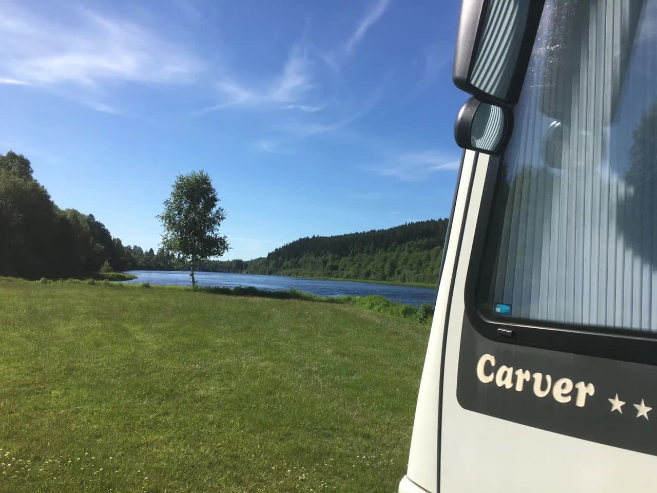En husbil står parkerad nära en sjö vid Värnäs Camping, omgiven av grönskande växtlighet. I bilens sidospegel reflekteras delar av det natursköna landskapet och sjön.