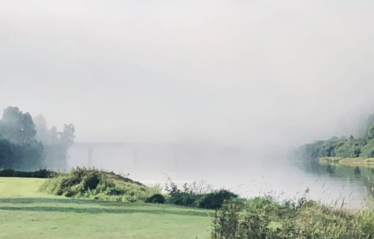En dimmig morgon vid en sjö eller flod, med tät dimma som svävar över vattnet och hindrar sikten, vilket skapar en lugn och mystisk atmosfär vid Värnäs camping.