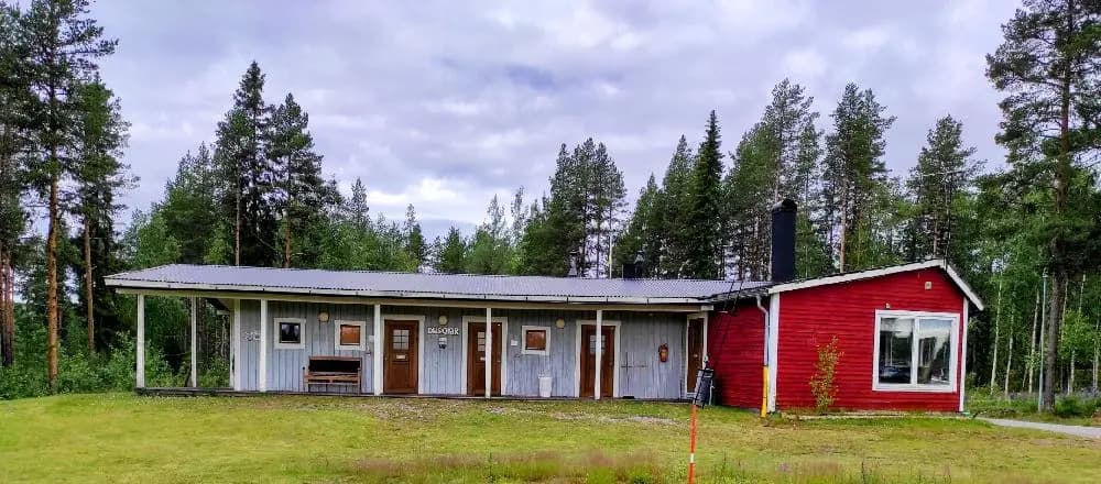 En mysig stuga omgiven av grönska på Meselefors camping, med en välskött gård och höga barrträd i bakgrunden.