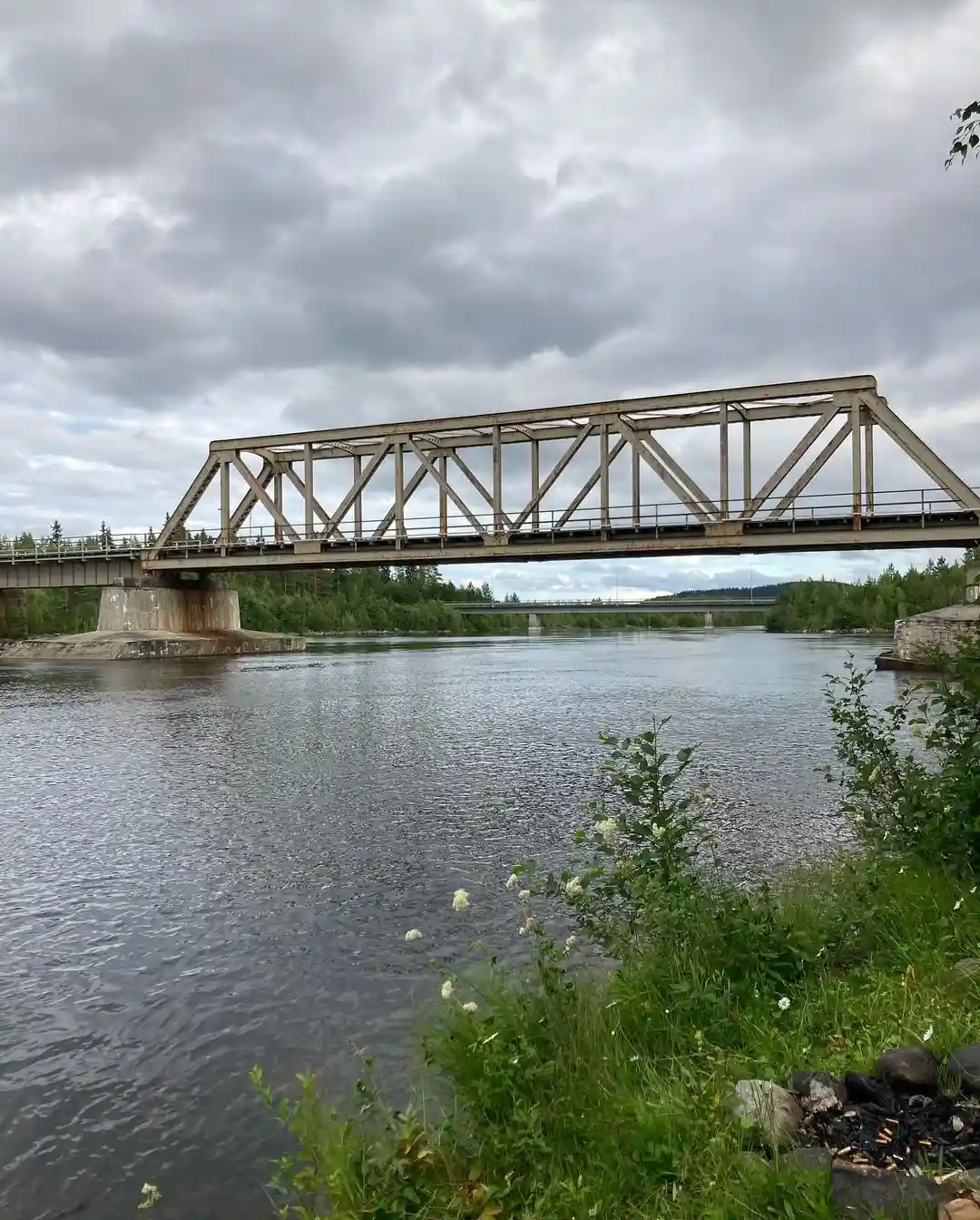 Bild av Meselefors camping med en bro som sträcker sig över en stilla flod. Moln täcker delvis den blå himlen, och omgivningen är grönskande med trädkantade flodbanker.