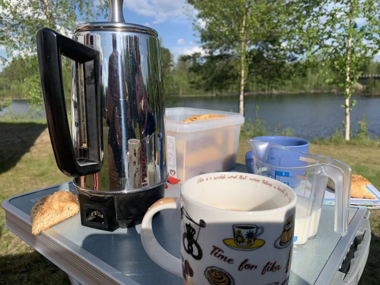 En kaffebryggare, en kopp och serveringskärl står på ett bord, omgivna av olika köksapparater och serveringstillbehör, typiska för en campingmiljö.