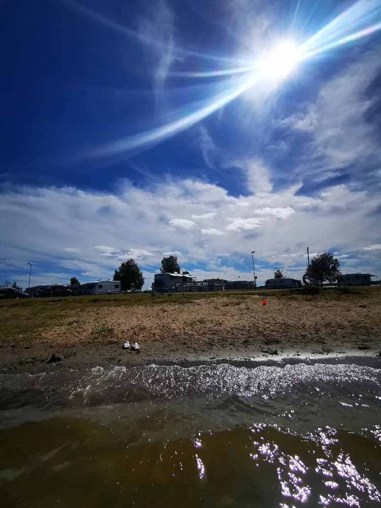 Bild av en klarblå himmel med vita, fluffiga cumulusmoln ovanför Borgaruddens havsbad och familjecamping, badplatsen sträcker ut sig mot horisonten. Solens strålar lyser upp den fridfulla miljön.